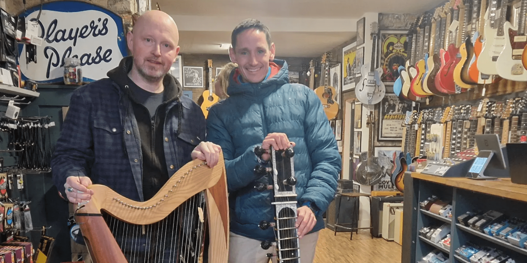 Two men posing with musical instruments, a harp and a sitar. There is a wall of guitars behind them.