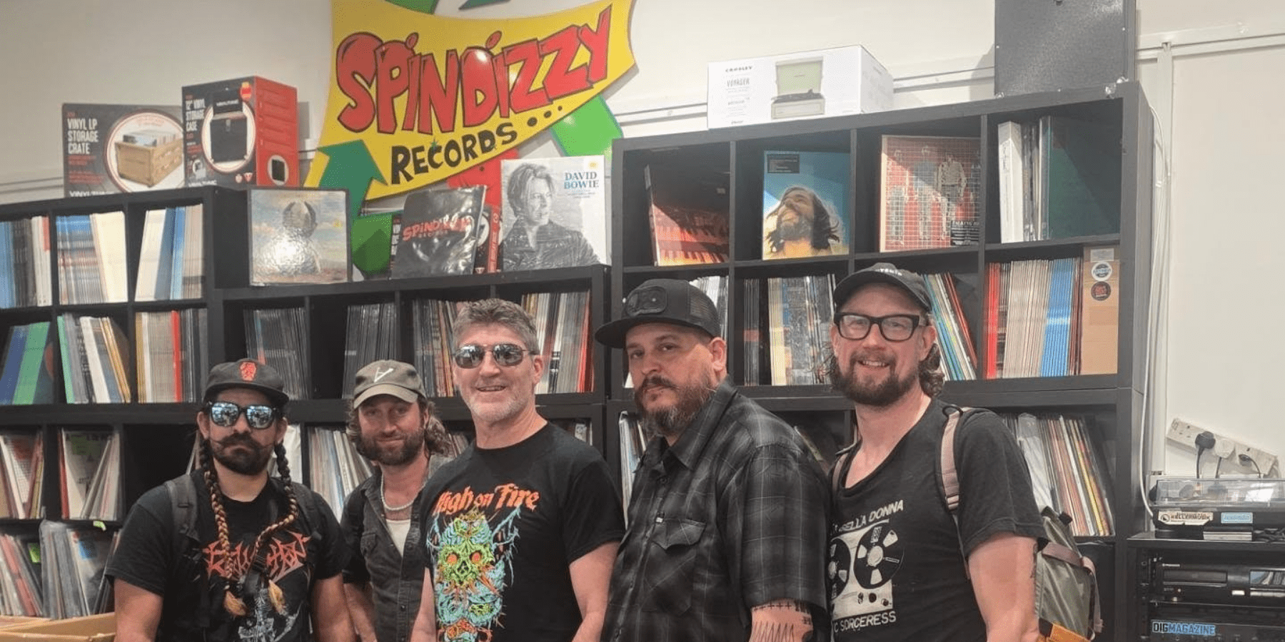 5 men posing in front of a large record collection. A spindizzy sign is visible above the record collection.