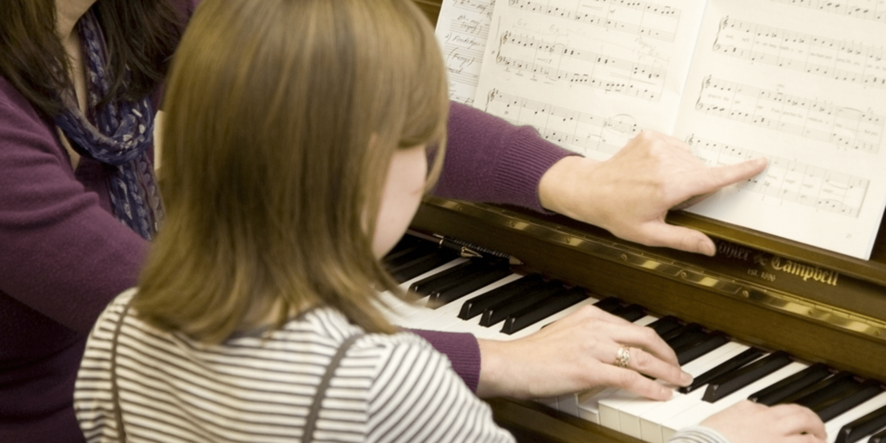 A woman and child sitting at a piano. The woman is pointing at the sheet musicon the piano.